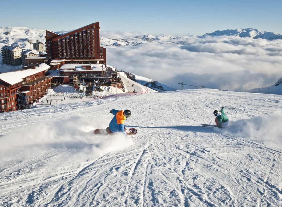 Dois snowboarders descendo a pista de Valle Nevado, com o hotel da estação à esquerda e um mar de nuvens sobre o vale ao fundo