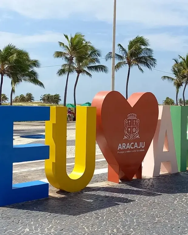 Letreiro colorido EU AMO ARACAJU na orla, com os arcos e coqueiros ao fundo